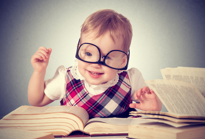 funny baby girl in glasses reading a book in a library